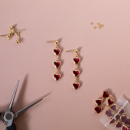 Heart-shaped earrings on a pink background with pliers and packaging.