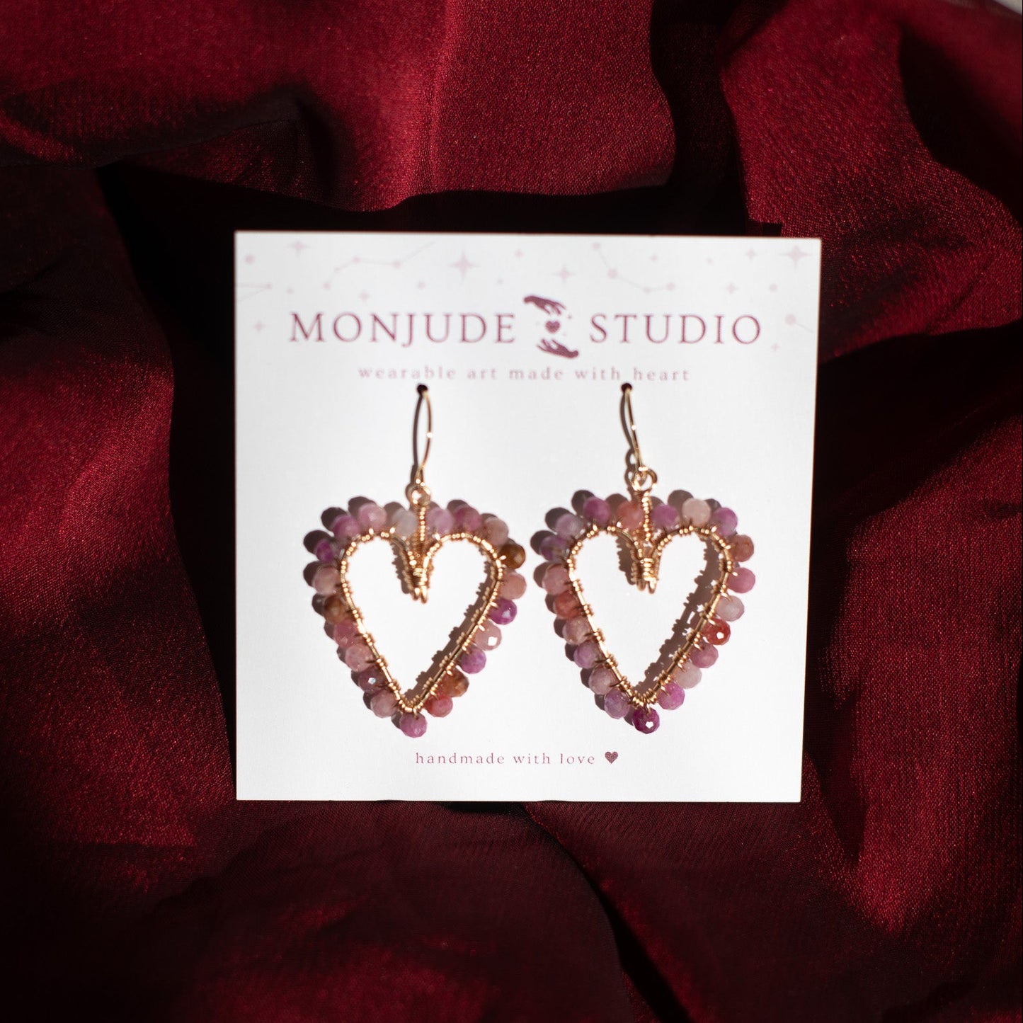 Heart-shaped earrings on a Monjude Studio card against a red fabric background