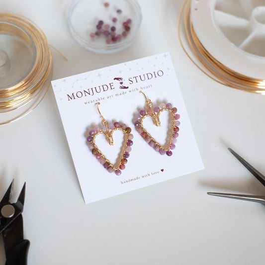 Heart-shaped earrings on a card with 'Monjude Studio' branding, surrounded by jewelry-making tools on a white surface.