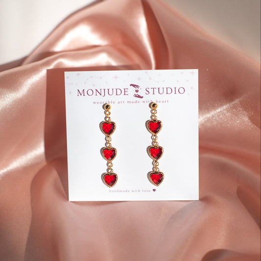 Heart-shaped earrings on a Monjude Studio card with pink fabric background