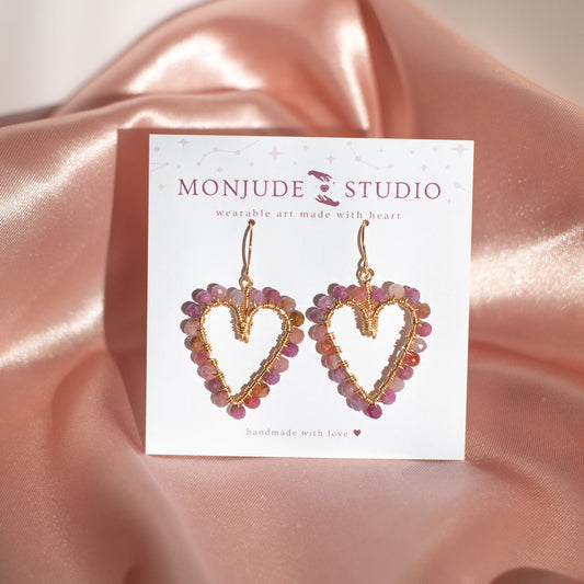 Heart-shaped earrings on a Monjude Studio card against a pink satin background
