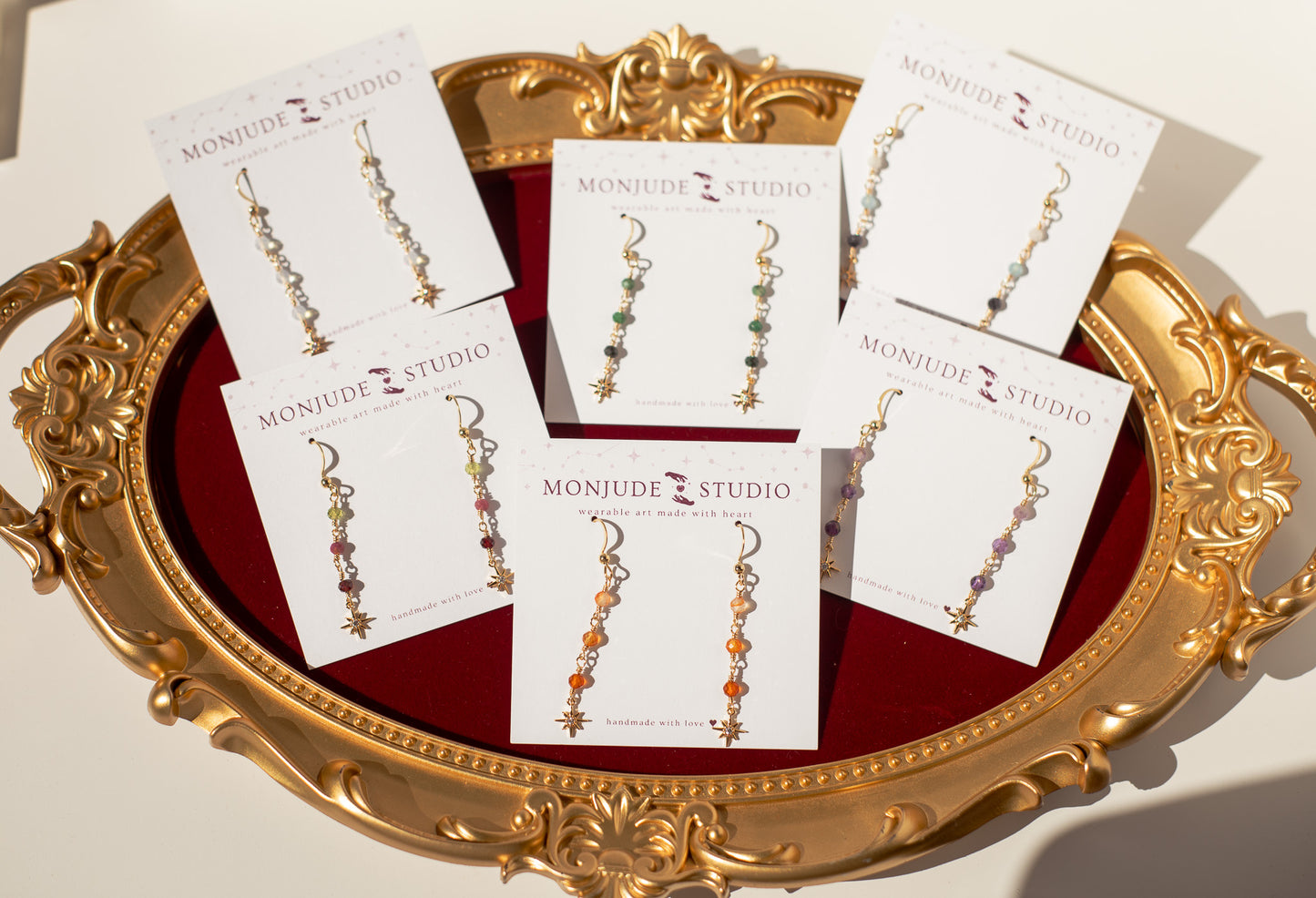 Six pairs of dangling earrings on white display cards branded MONJUDE STUDIO, arranged on an ornate gold tray with a red velvet lining.
