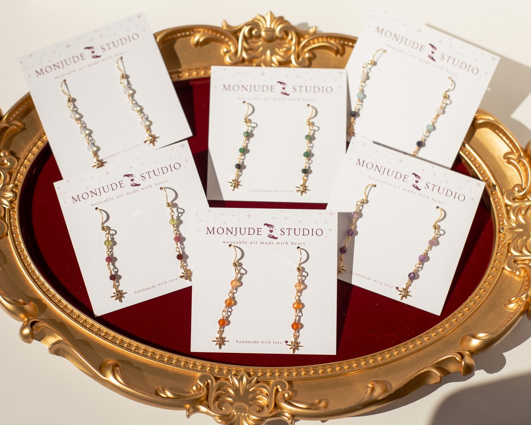Six pairs of dangling earrings on white display cards branded MONJUDE STUDIO, arranged on an ornate gold tray with a red velvet lining.
