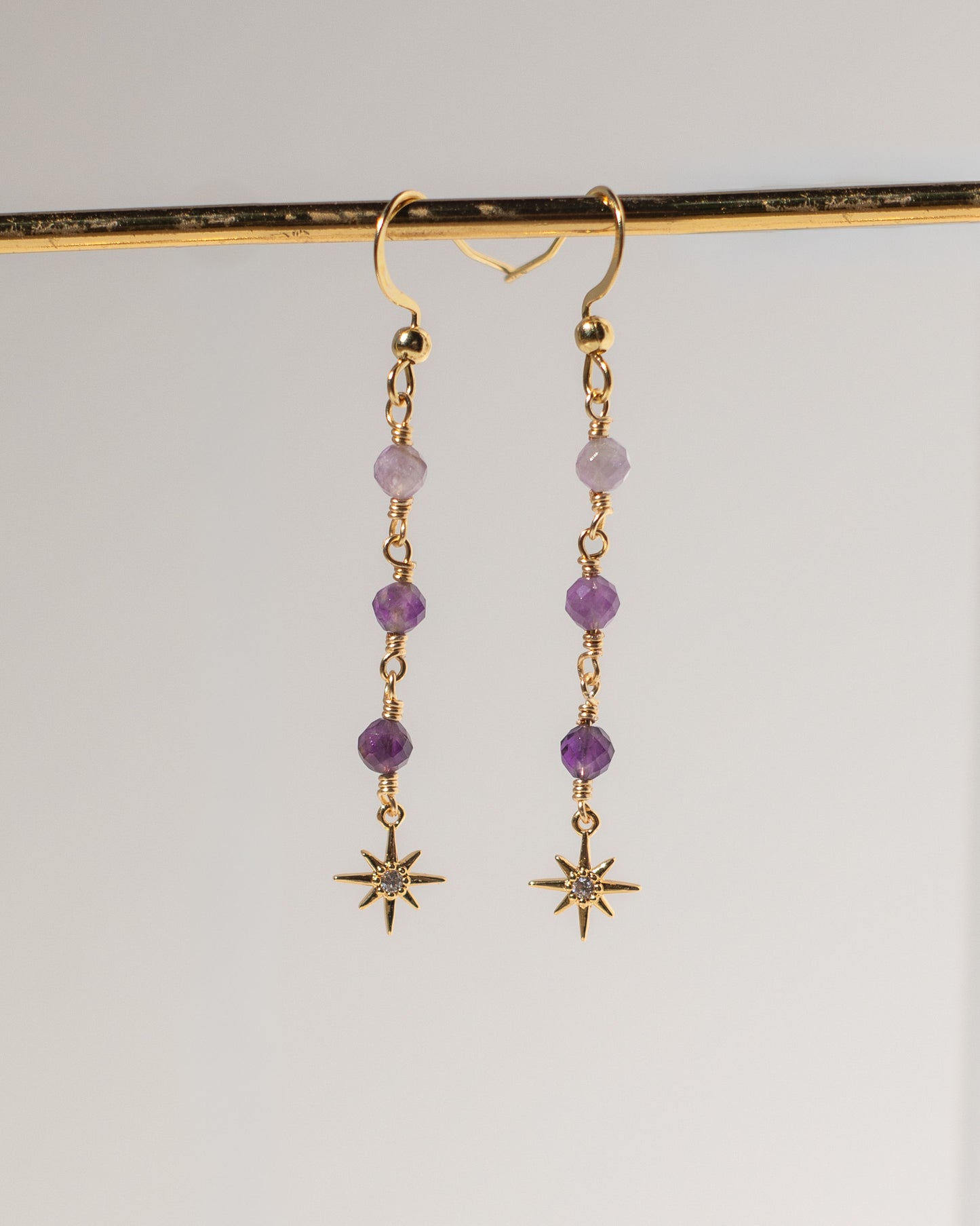 A pair of gold dangle earrings with purple beads and starburst charms, hanging from a gold display bar against a light gray background.