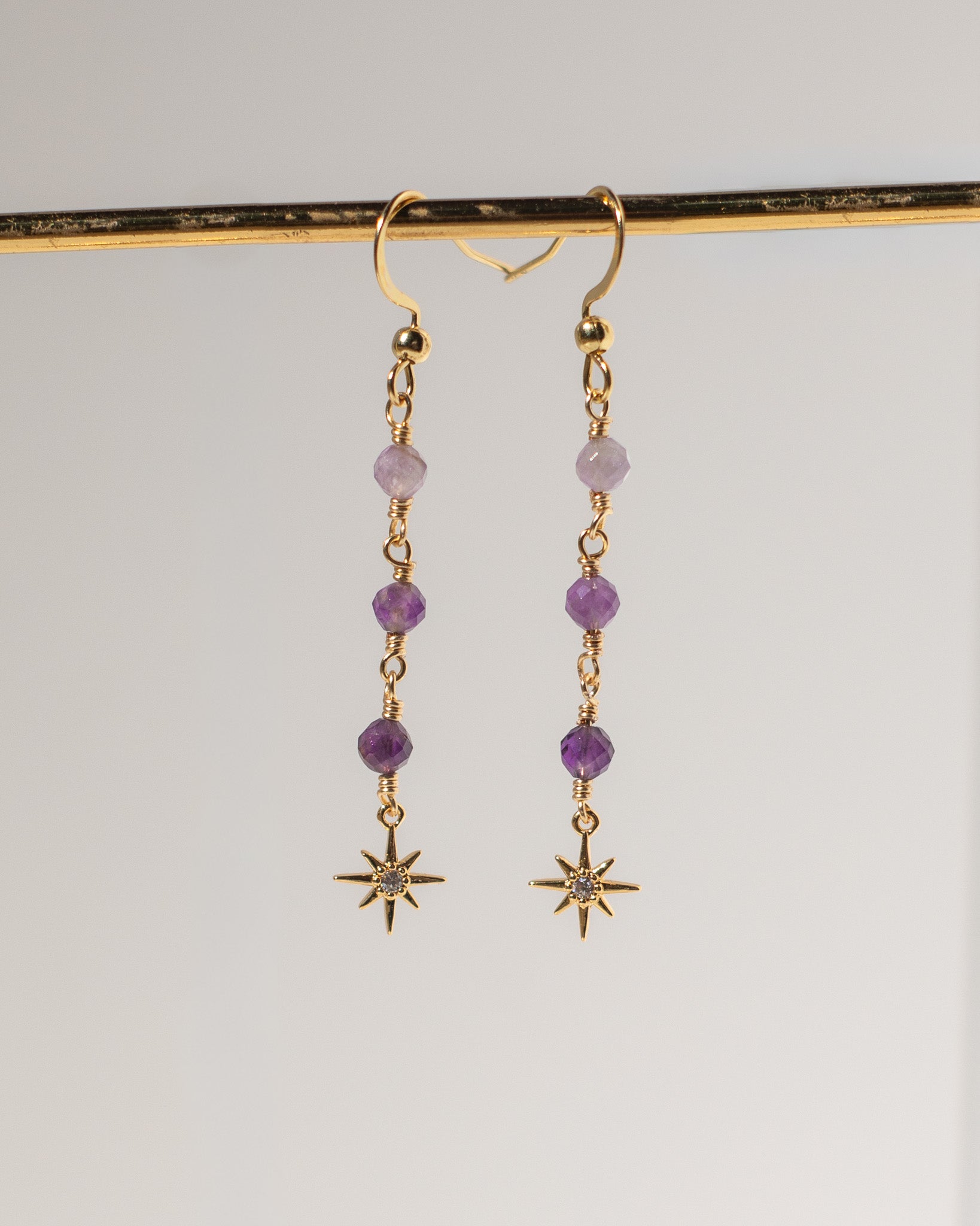 A pair of gold dangle earrings with purple beads and starburst charms, hanging from a gold display bar against a light gray background.