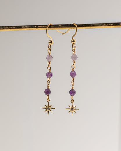 A pair of gold dangle earrings with purple beads and starburst charms, hanging from a gold display bar against a light gray background.