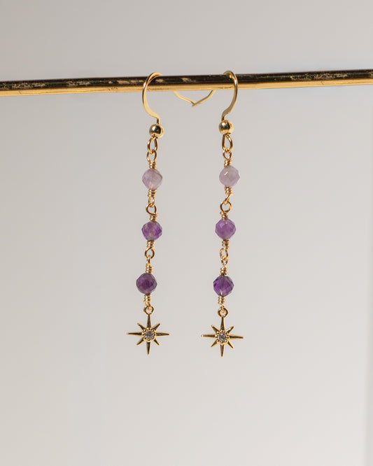 A pair of gold dangle earrings with purple beads and starburst charms, hanging from a gold display bar against a light gray background.